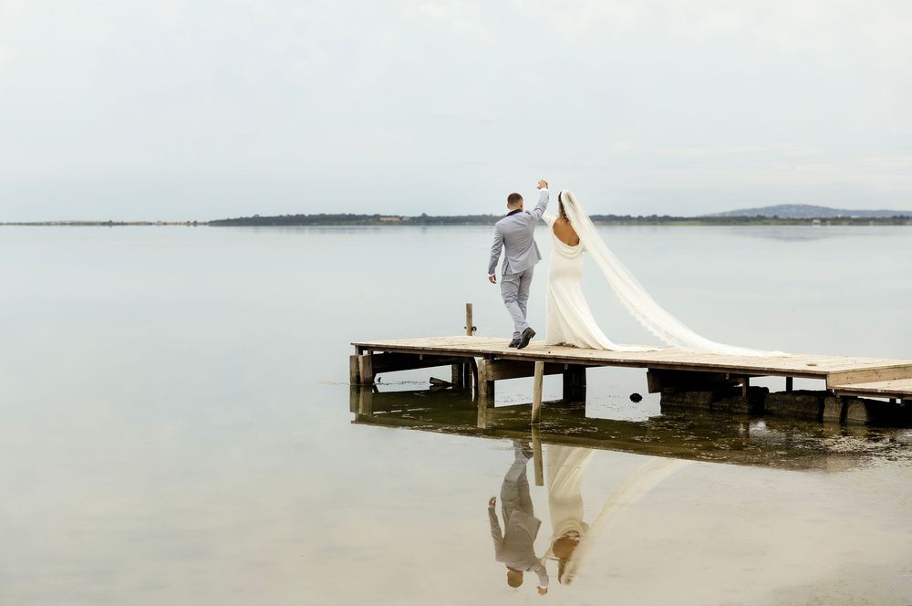 Bride and groom walking on wooden dock over calm lake, wedding dress veil flowing in the wind.