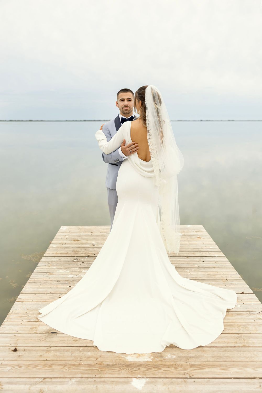Bride and groom embrace on a wooden dock overlooking calm water, bride wearing elegant white gown with veil and train.