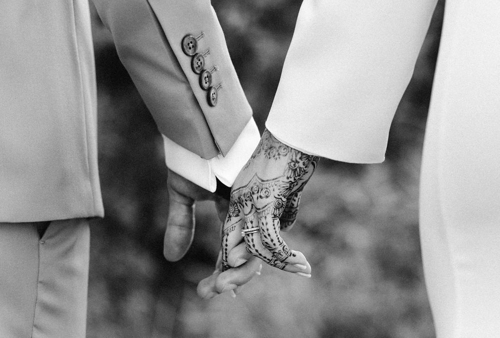 Bride and groom holding hands, showcasing intricate henna mehndi design on bride's hand, black and white wedding photo.