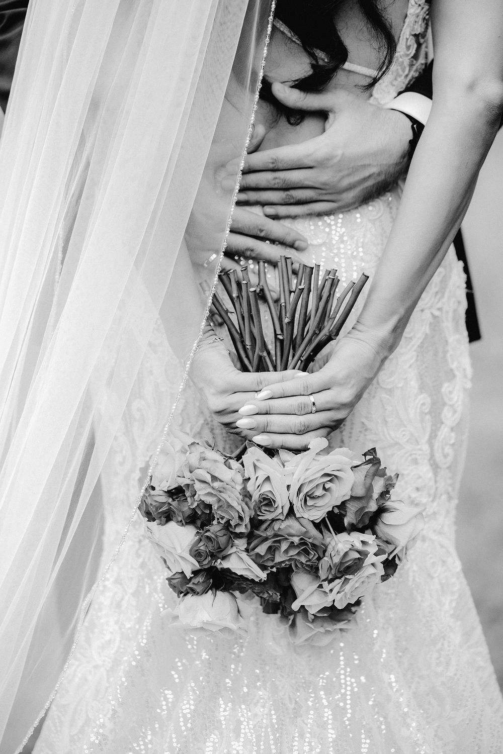 Black and white photo of bride and groom embracing, bride holding a bouquet of roses in a lace wedding dress.