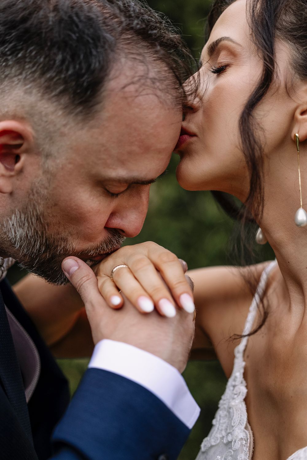 Groom kisses bride's hand while she kisses his forehead during romantic outdoor wedding ceremony.