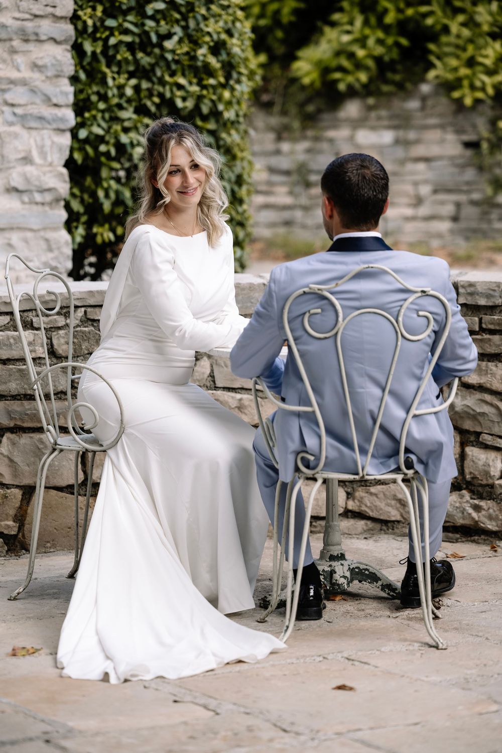 Bride in white gown and groom in light blue suit sit on ornate garden chairs, holding hands at outdoor wedding venue.