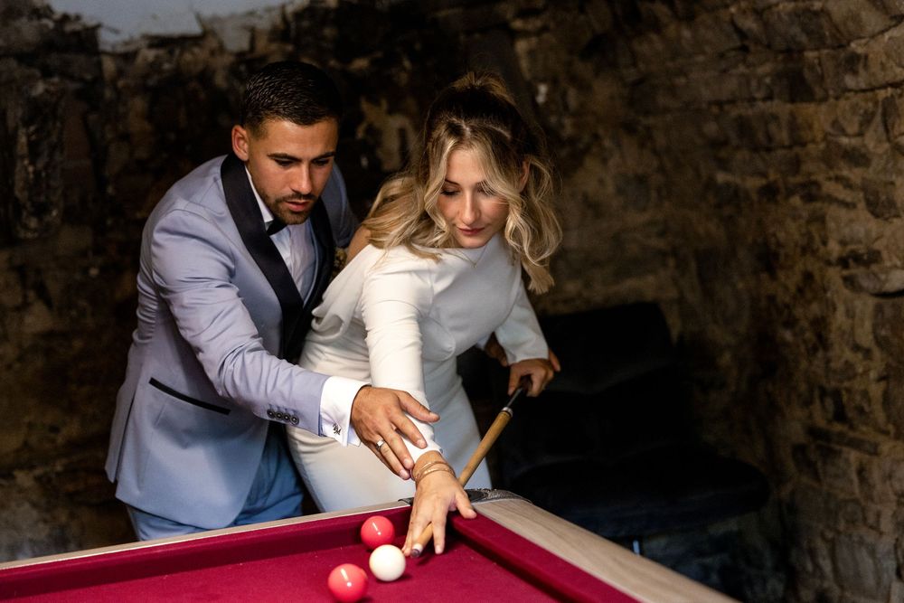 Elegant couple in formal attire playing pool together on a red billiard table in a rustic stone setting.