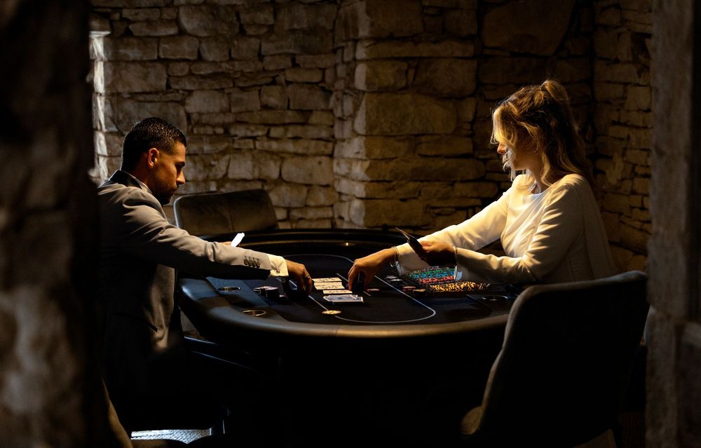 Two people playing cards at a dark casino poker table in a dimly lit stone room.