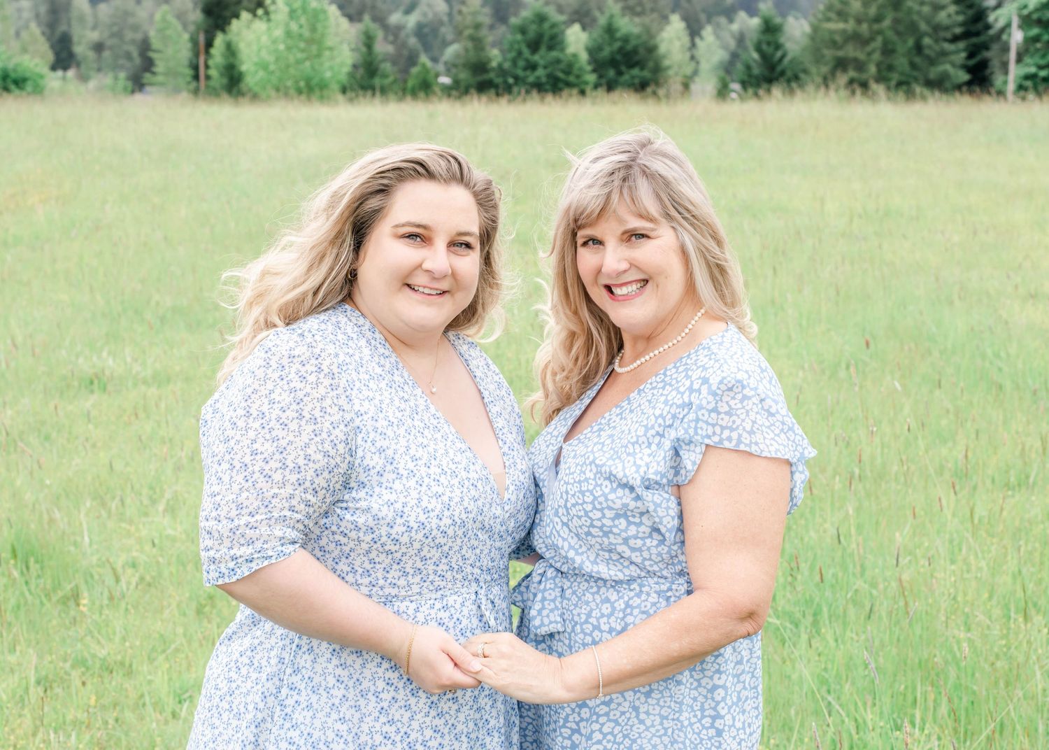 Two people in matching light blue floral dresses pose together in a scenic mountain meadow.