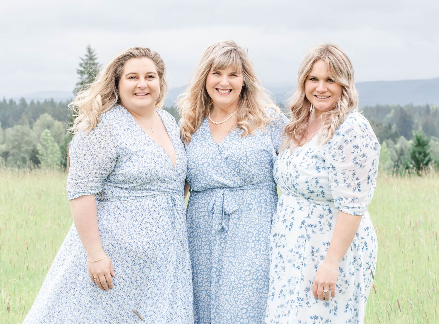 Three people wear coordinating blue floral dresses while posing in a scenic mountain field.