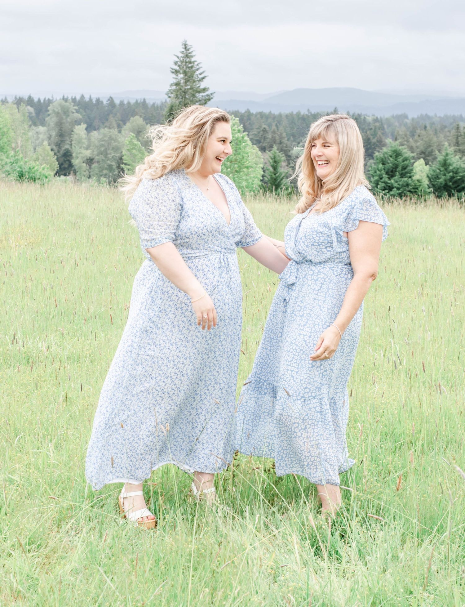 Two people in flowing blue dresses share a laugh in a sunlit mountain meadow.