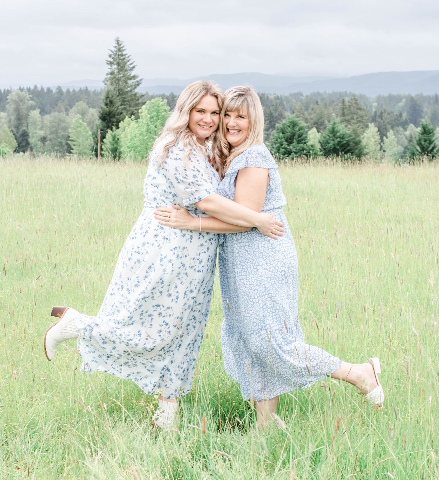 Two people in floral dresses embrace joyfully in a green mountain meadow.