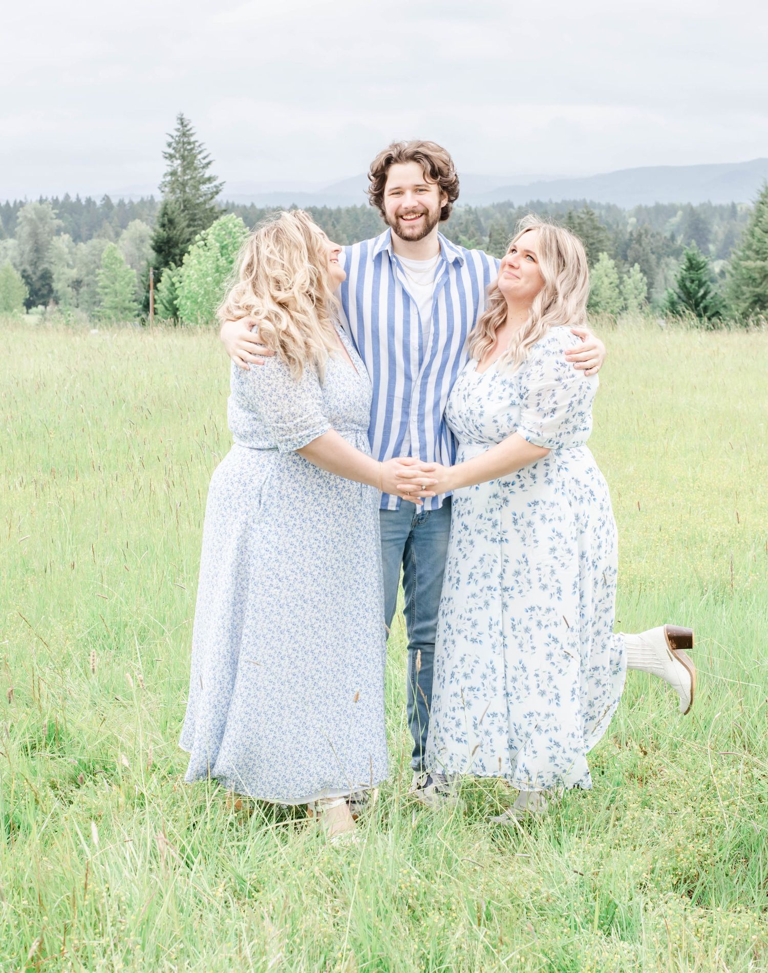 Three people in coordinated light outfits pose together in a sunny meadow with mountains and evergreen trees in background.