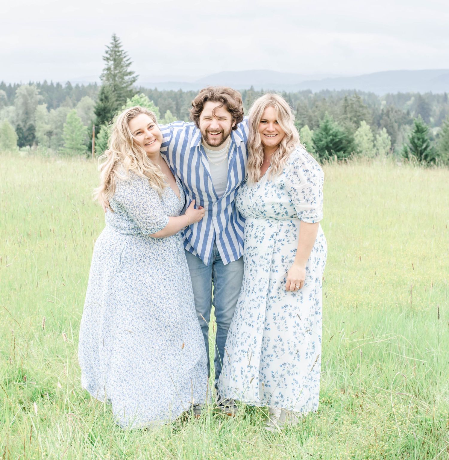 Three people in white and blue outfits embrace while standing in a grassy field with forest views.