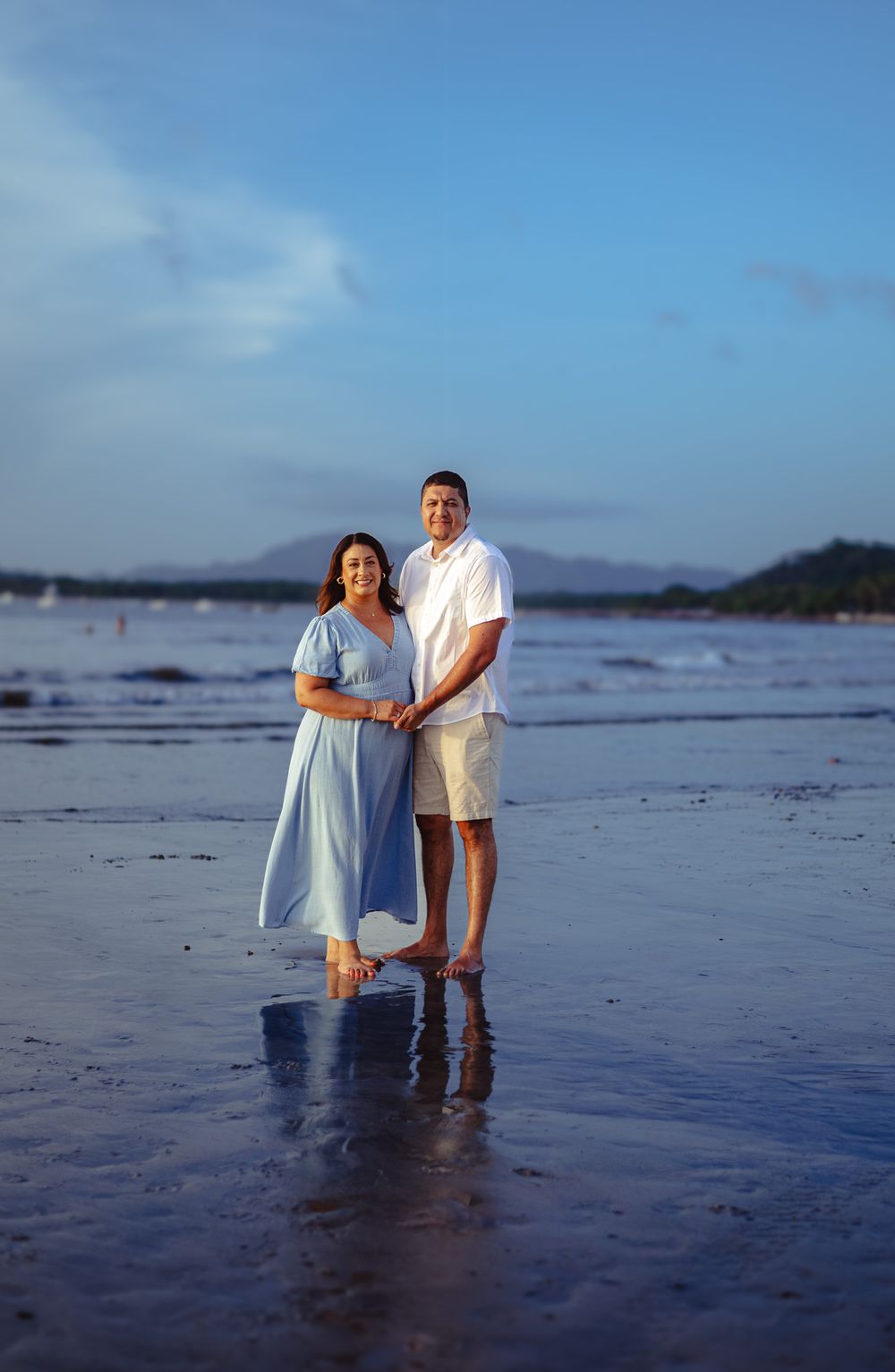 A couple stands together on a beach at dusk with mountains in the background.