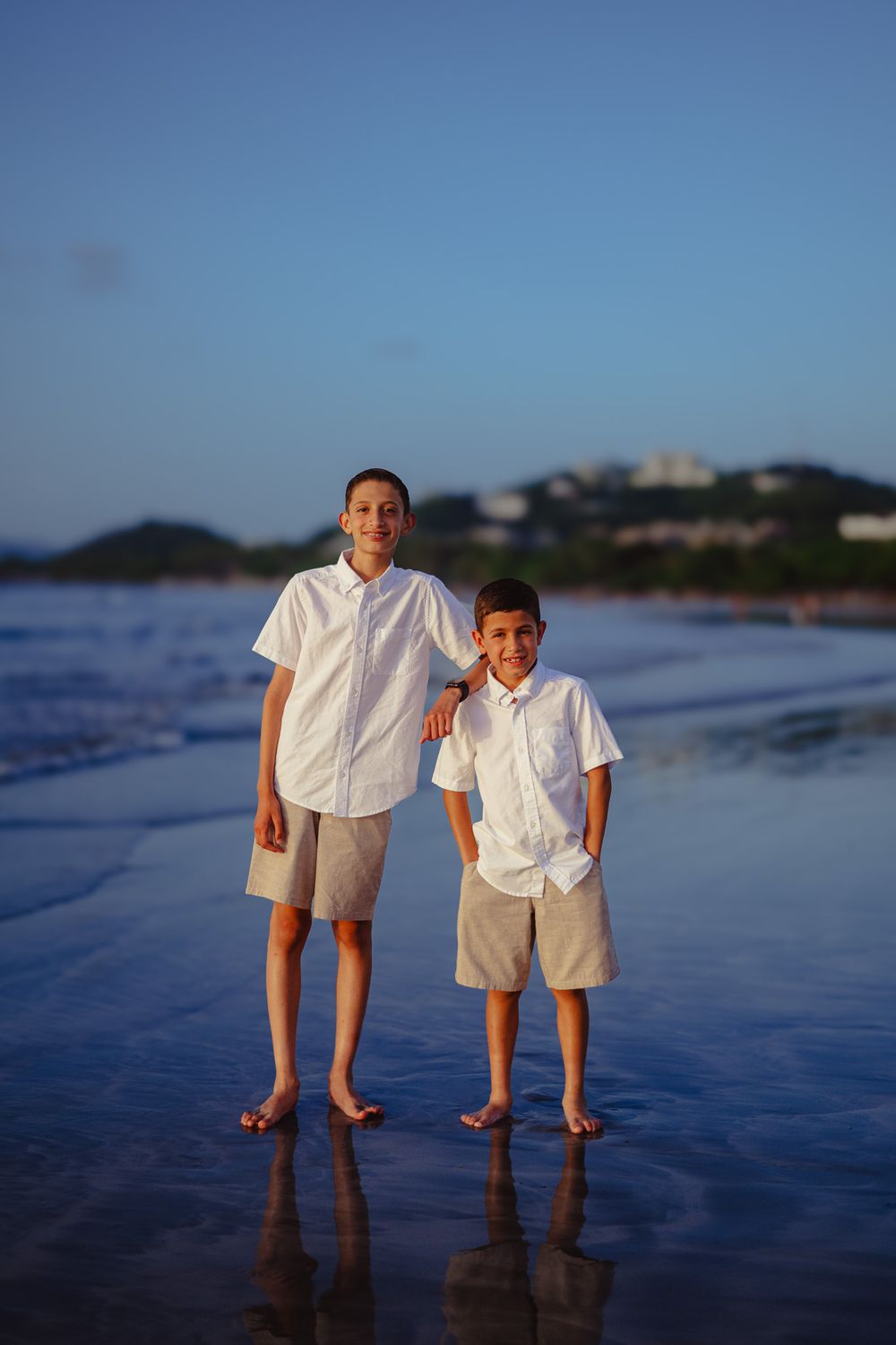 Two people in matching white shirts stand together on a wet beach at sunset.