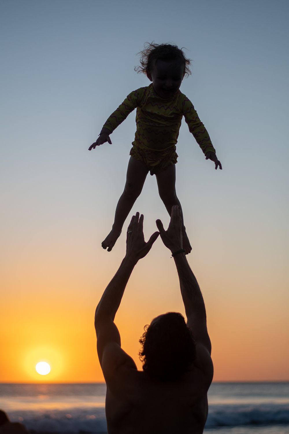 Silhouette of parent tossing child in air against sunset beach backdrop.
