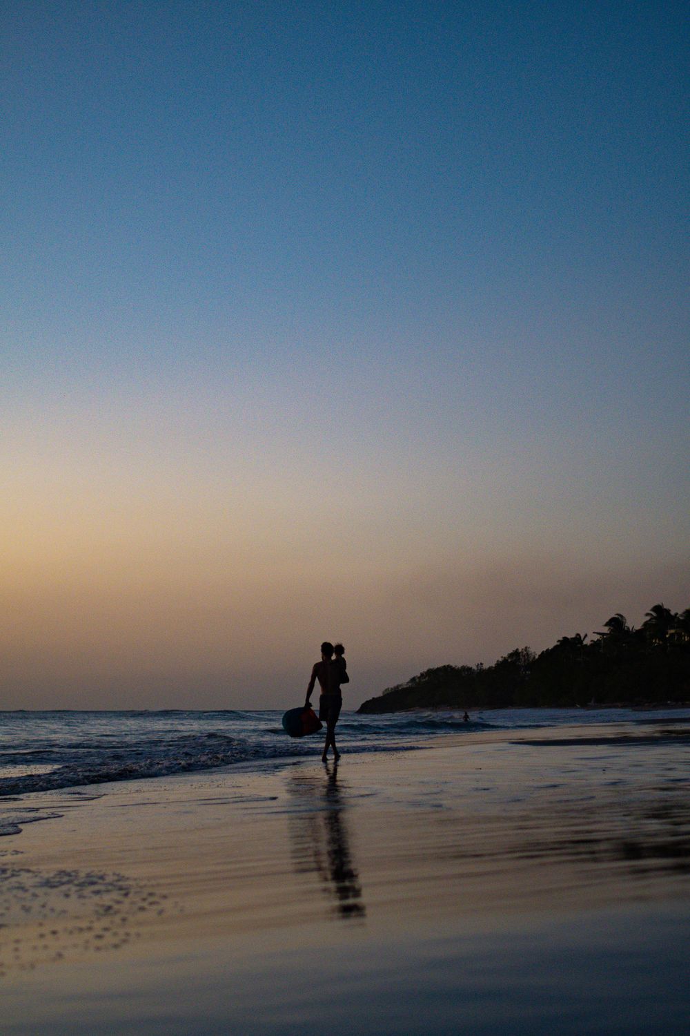 Lone figure walking on beach at dusk with surfboard carrying silhouetted against sunset sky.