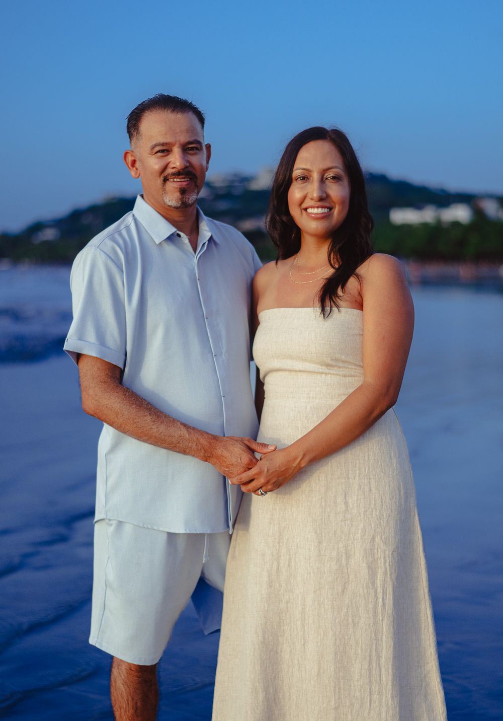 A couple in white beach attire poses together on a sandy shore at dusk.