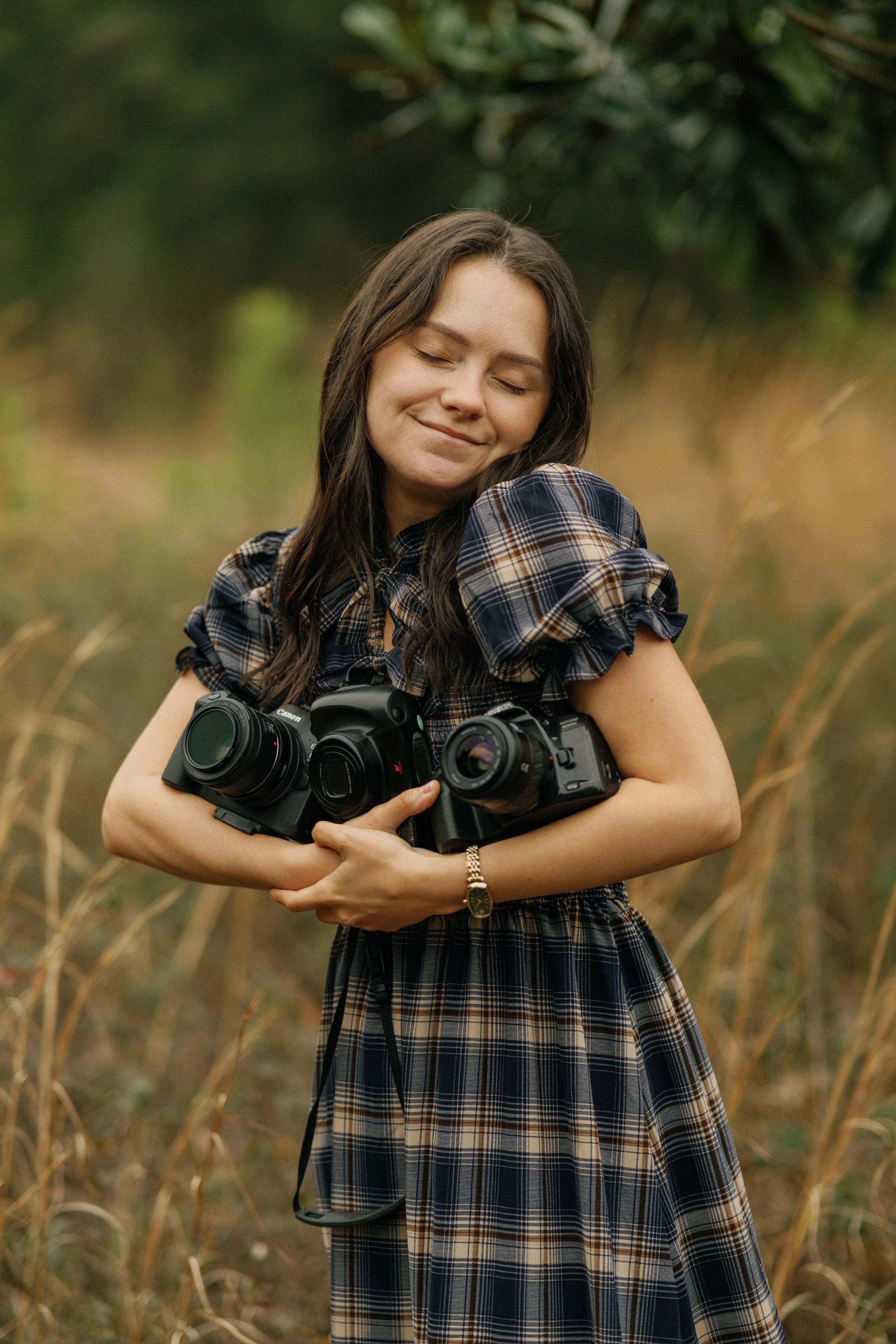 A smiling young woman in a plaid dress hugs multiple cameras while standing in a golden field of tall grass.