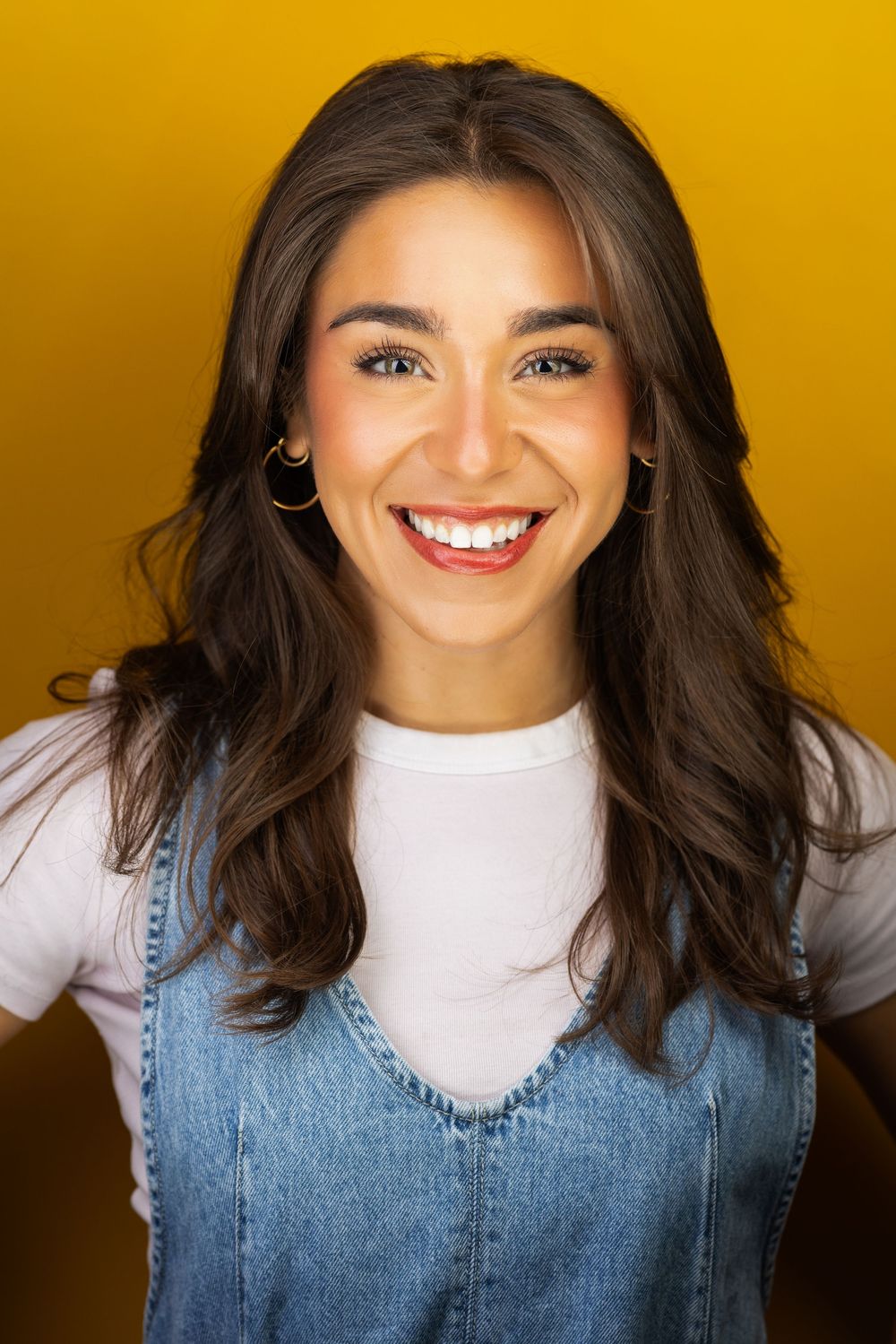 Young smiling brunette woman in denim vest and white shirt against yellow background.