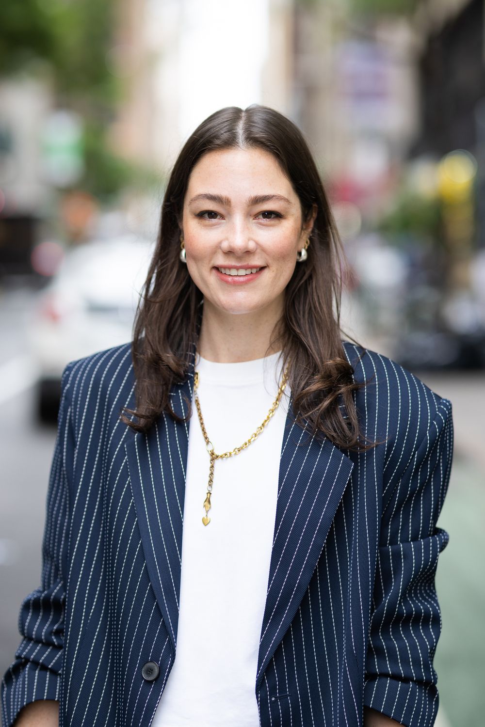 Smiling brunette woman in navy pinstripe blazer with gold necklace on city street.