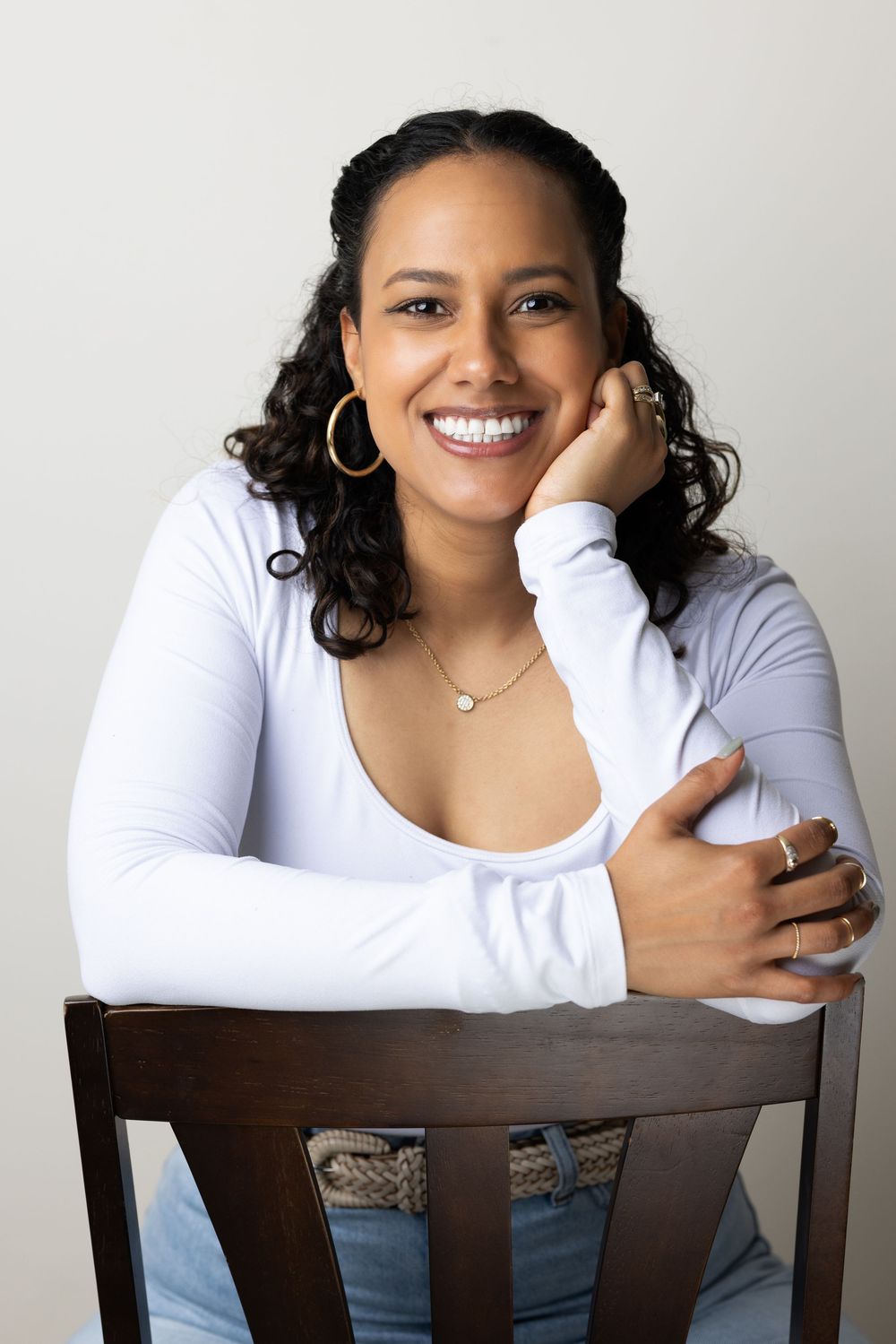 Smiling woman with curly hair in white top seated on wooden chair against white background.