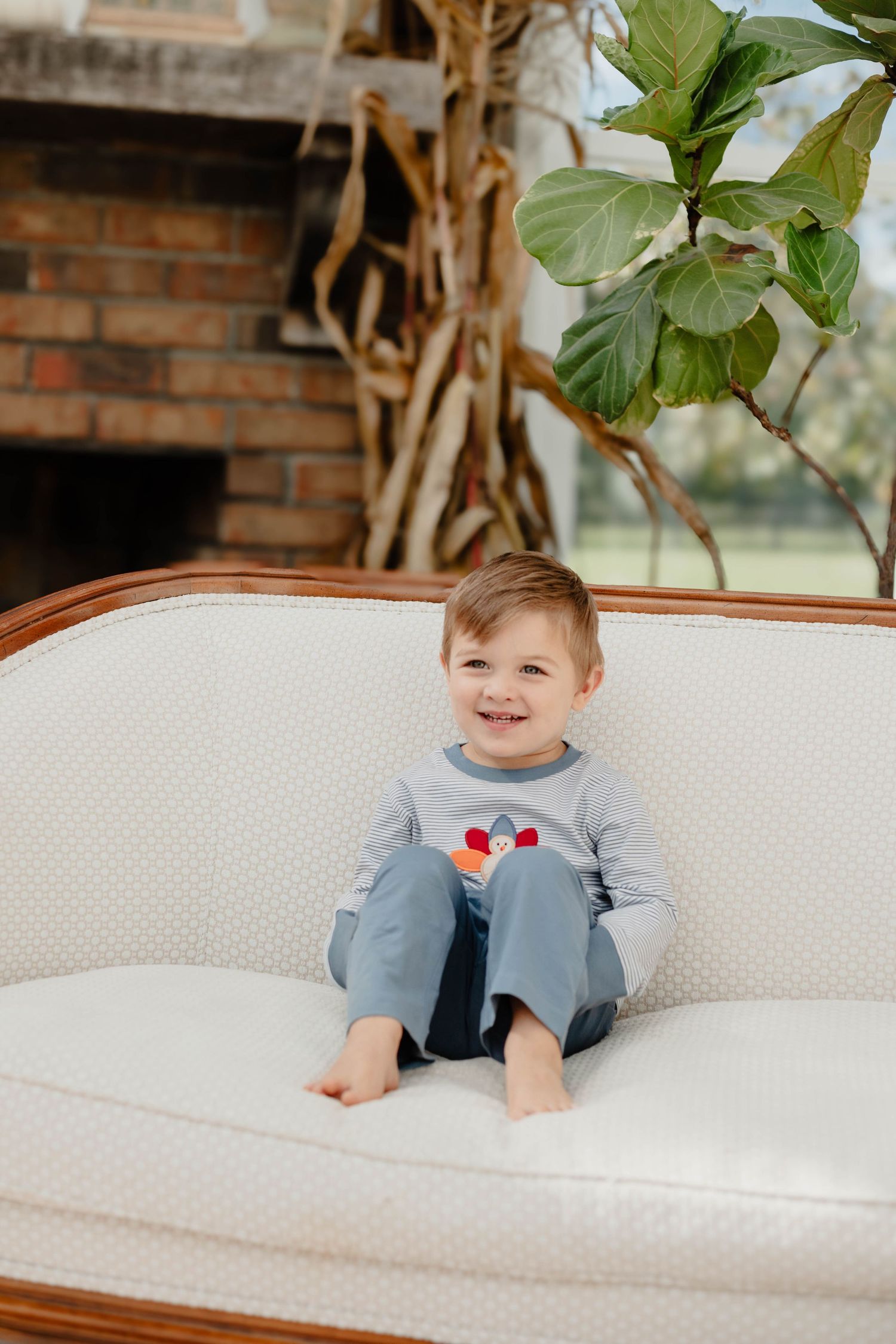 A small child sits barefoot on a white vintage sofa near a fiddle leaf fig plant.