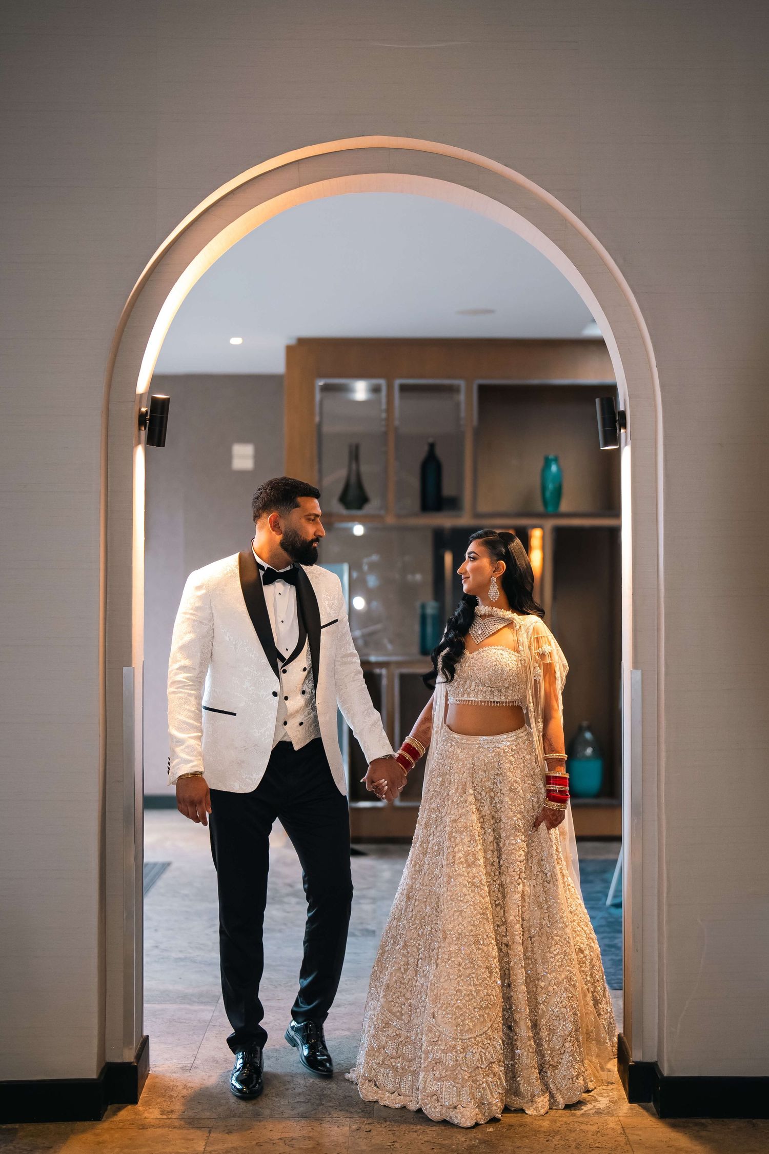 Couple in elegant wedding attire holding hands under a glowing arch, groom in white tuxedo, bride in gold lehenga.