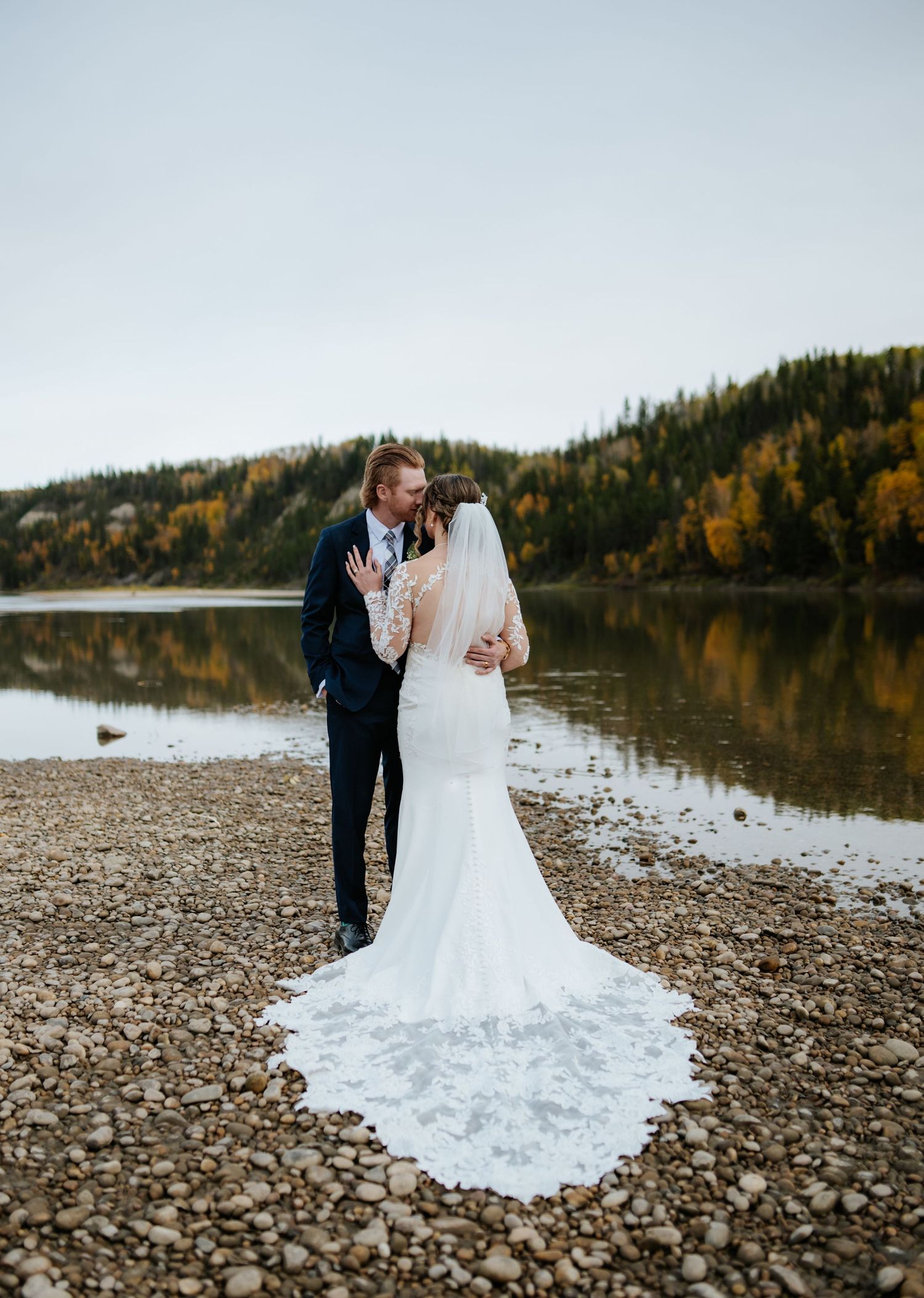 Bride in a lace gown with a long veil embraces her groom in a navy suit on a pebbly riverbank with autumn trees behind.
