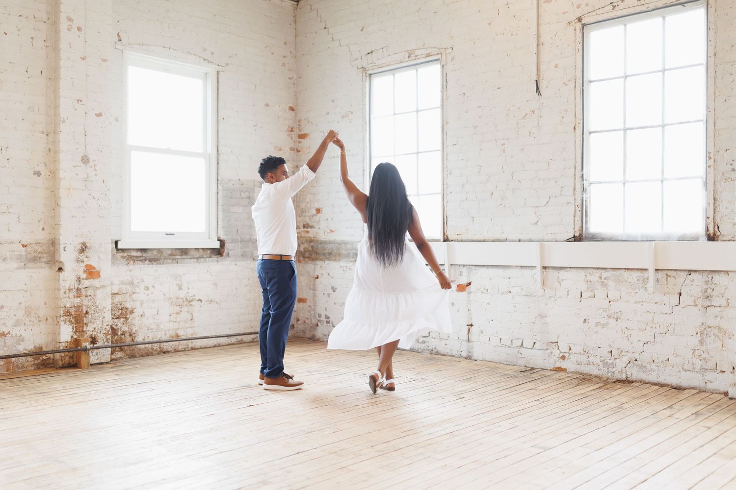 A couple dances romantically in an empty white room with sunlit windows.