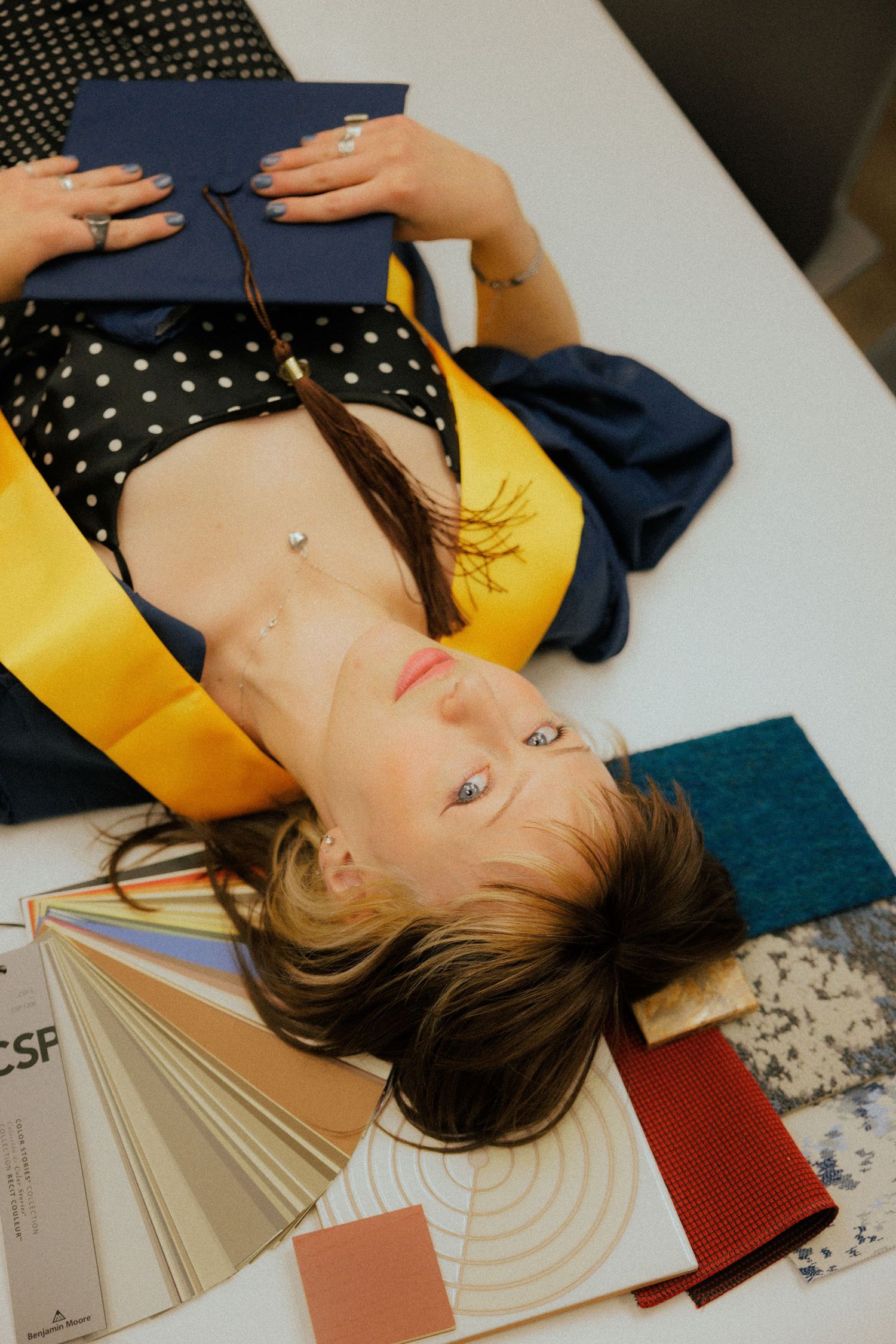 An interior design student lies on a white table surrounded by fabric swatches, clutching a graduation cap.