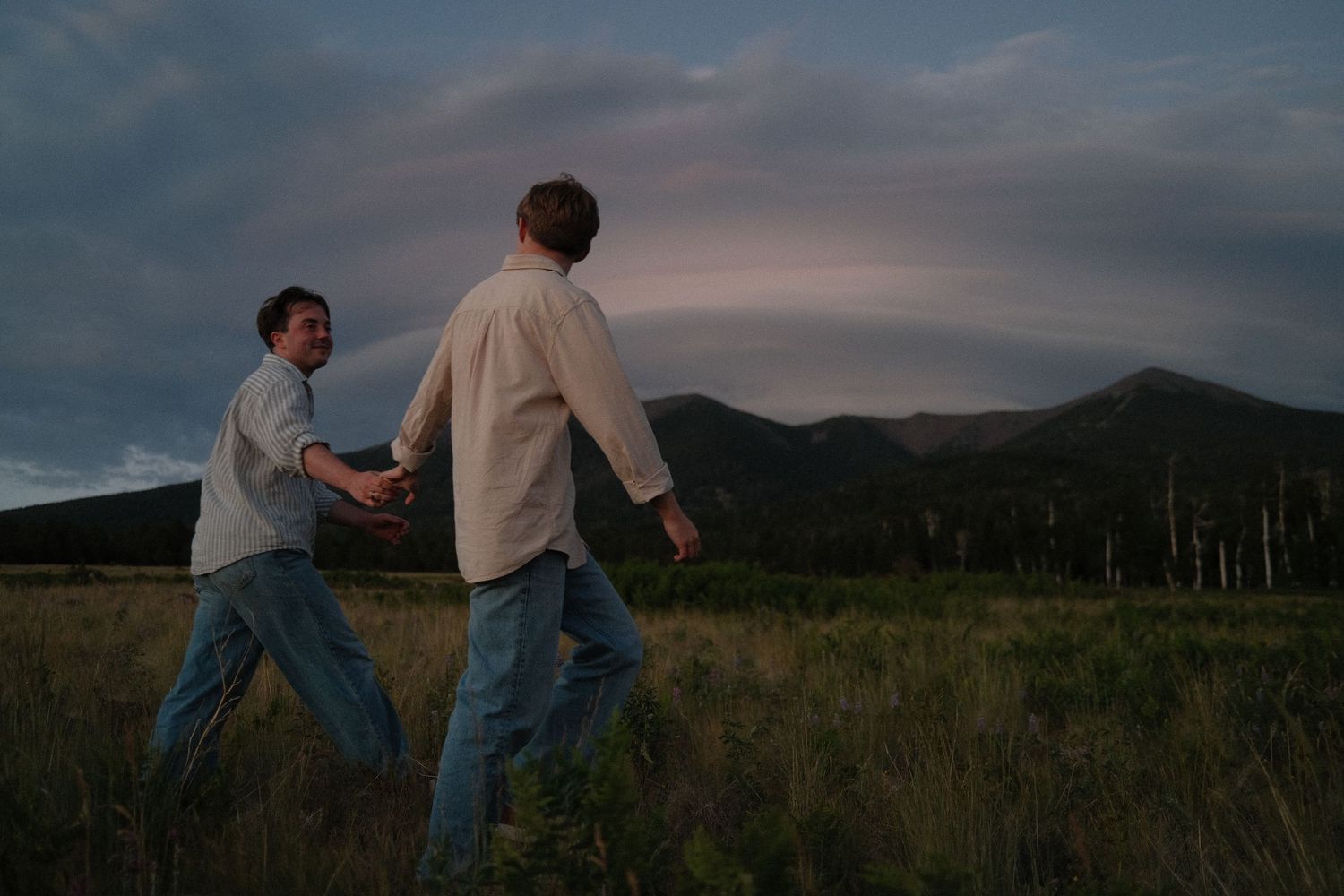 Two men hold hands while walking through a grassy mountain field at dusk under dramatic clouds.