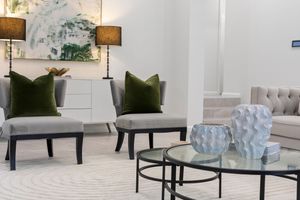 Modern living room with grey accent chairs, olive green pillows, and a glass coffee table on a light area rug.