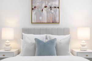 Modern bedroom interior with white bedding, blue accent pillows, and abstract art on the wall above the headboard.