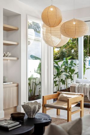 Modern dining area with wooden bench seating, paper lantern lights, and tall potted plants by large windows.