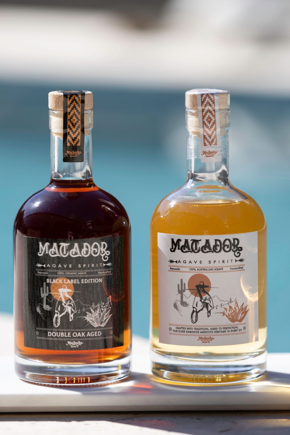 Two bottles of Matador spirits displayed against a blurred background, one dark amber and one light colored.