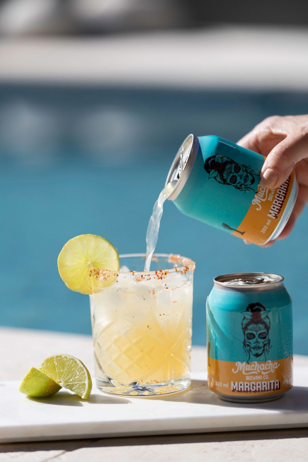Turquoise canned cocktail being poured into glass with lime garnish by pool.