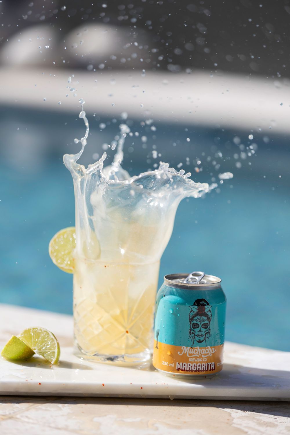 A can of margarita splashes into a glass with lime garnish by a pool creating dynamic water droplets against a blue background.