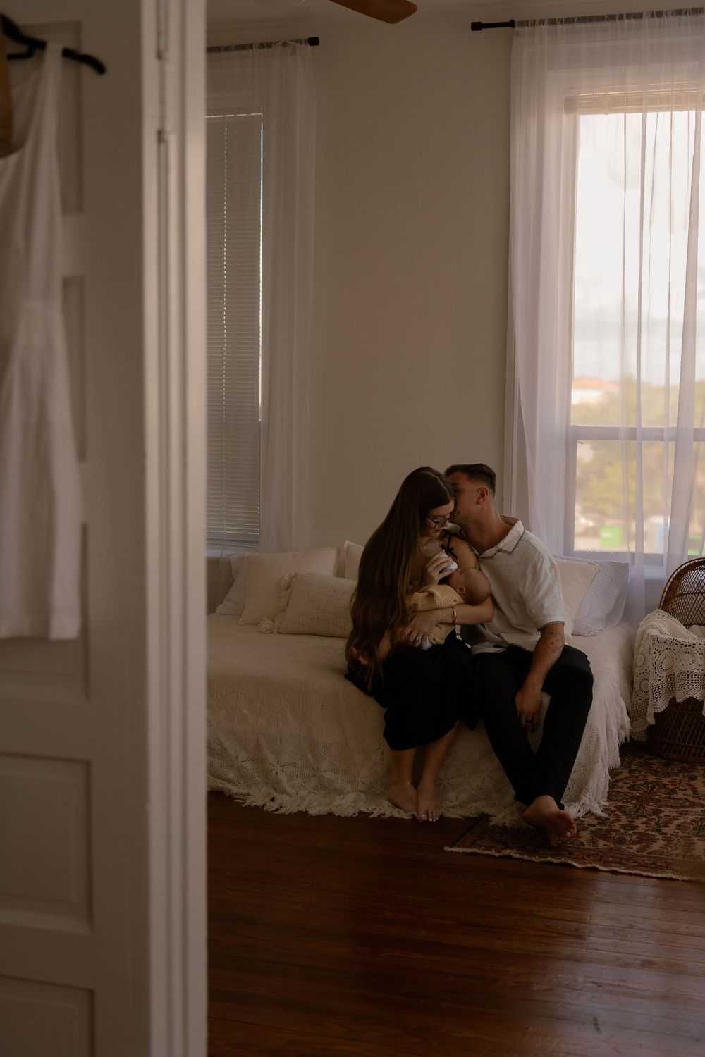 Couple sitting on bed sharing tender moment with newborn baby in softly lit bedroom.