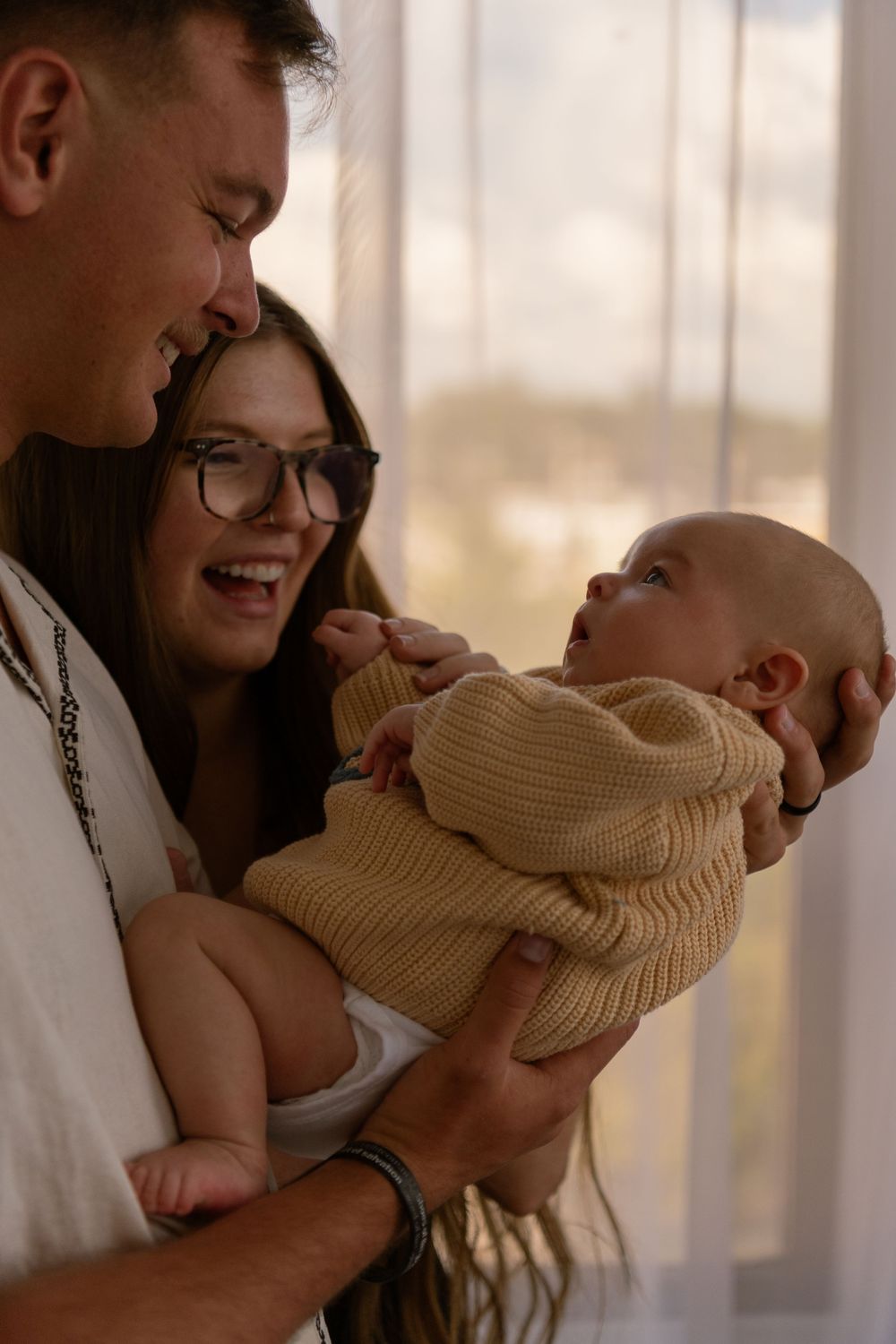 Smiling parents holding newborn baby in knit outfit near bright window.