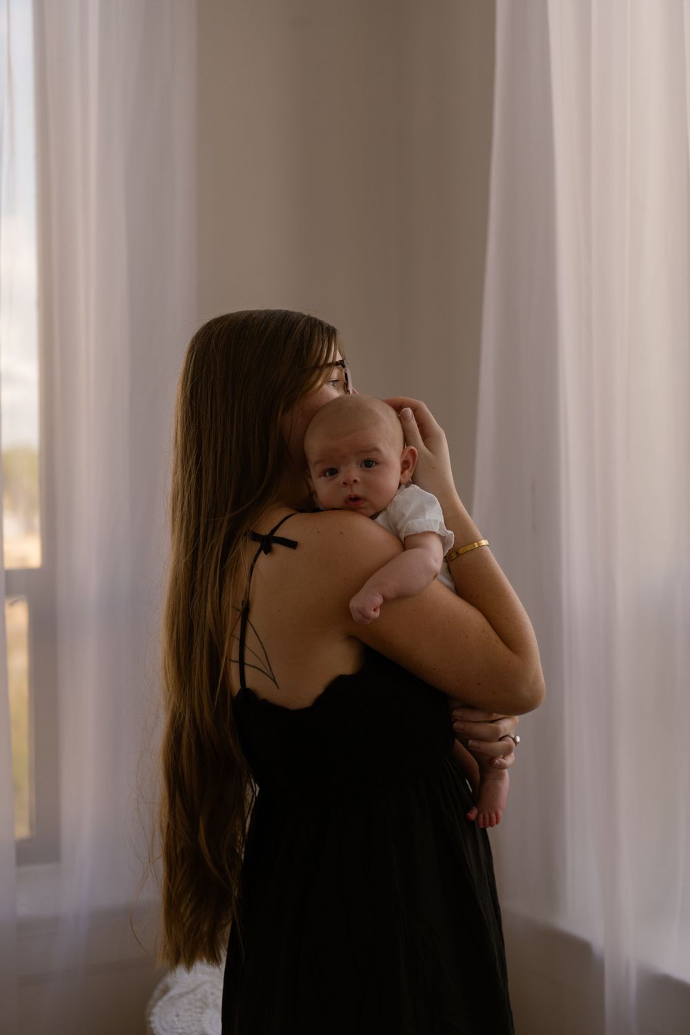 Mother with long hair holding newborn baby against her shoulder by sheer curtains.