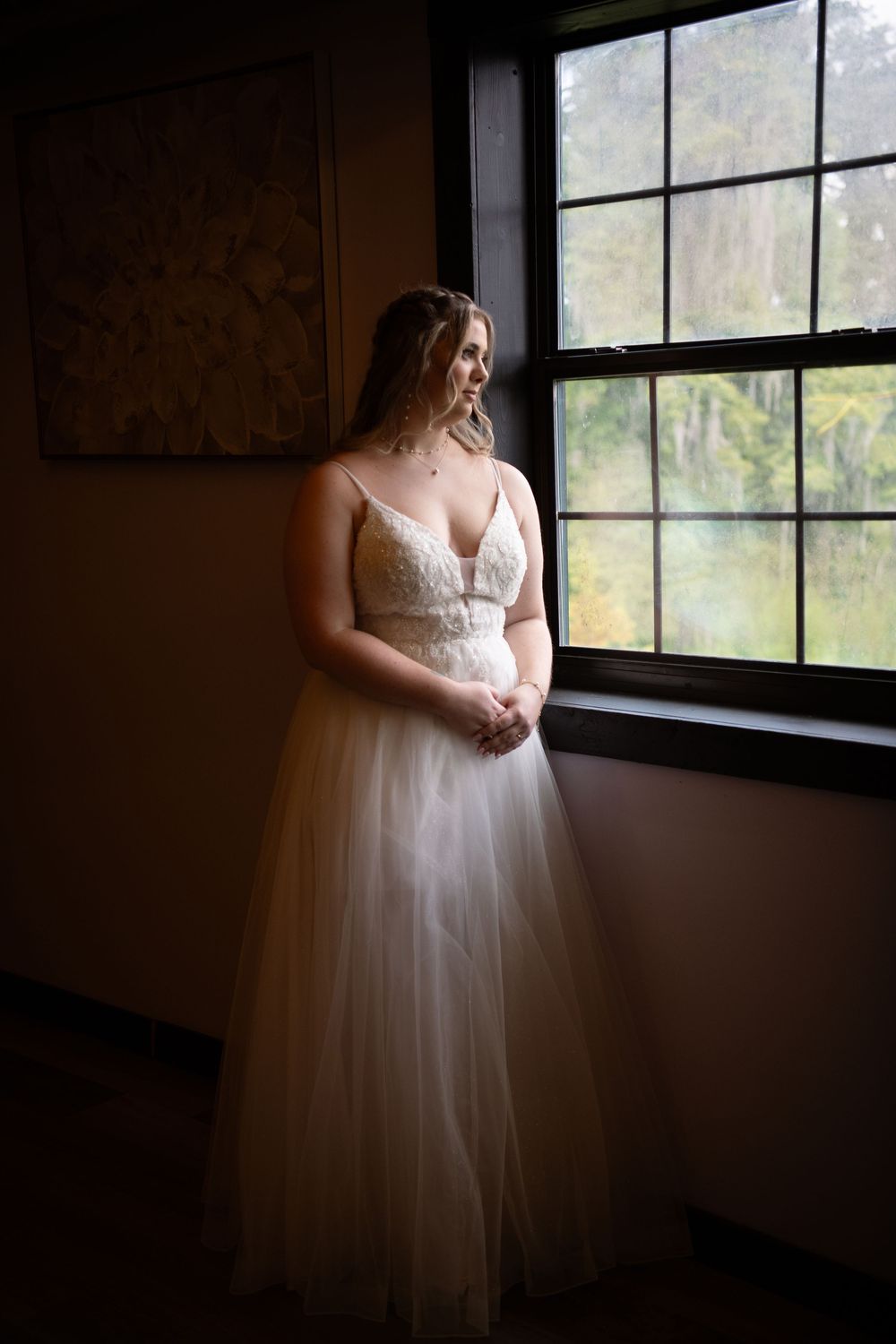 Bride in lace tulle gown standing by large window in soft natural light.