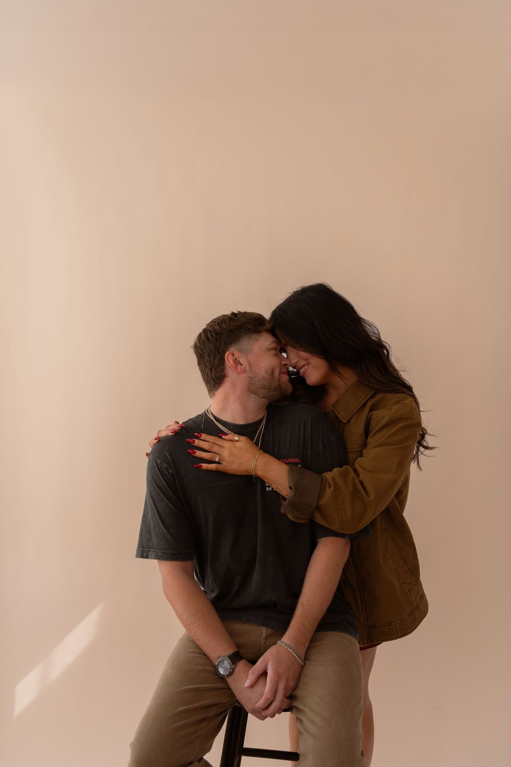 Romantic couple embracing in studio, woman in olive jacket resting forehead against seated man in gray tee.
