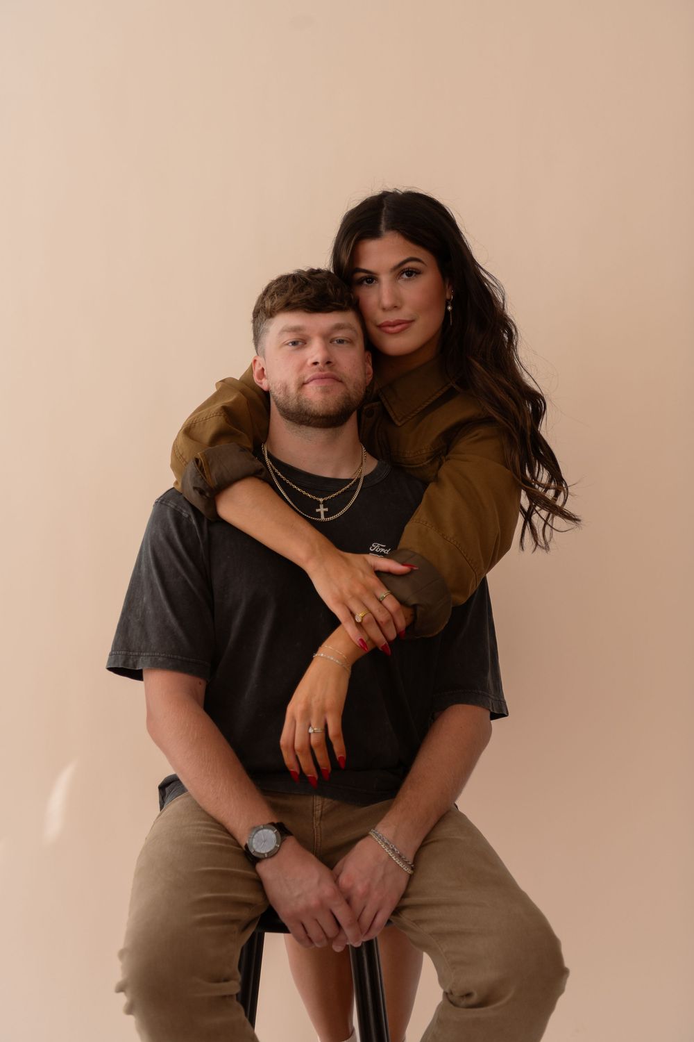 Couple posing in studio, woman in olive jacket hugging man from behind as he sits on stool.
