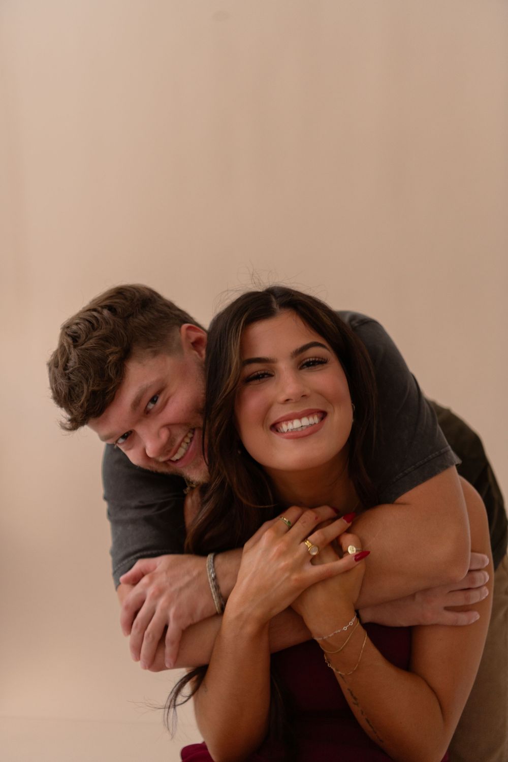 Joyful couple laughing as man hugs woman from behind, both smiling in a warm studio setting.