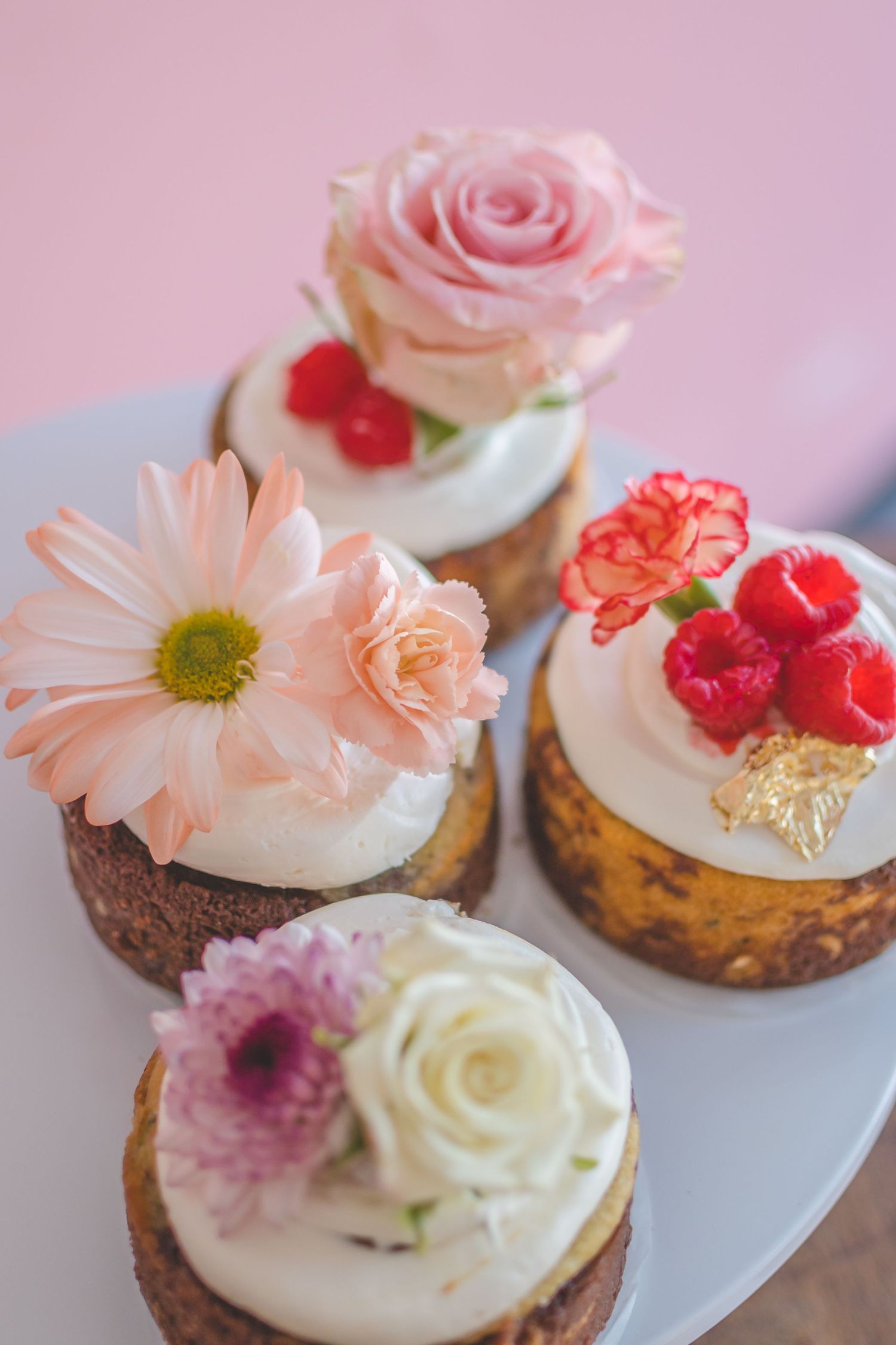 Gourmet cupcakes decorated with fresh flowers and berries on white plate against pink background.