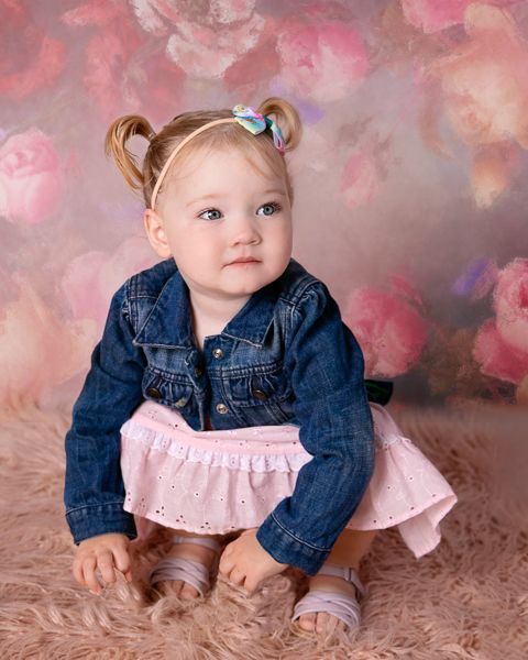 Adorable toddler girl in denim jacket and pink skirt with pigtails sits on fluffy rug against floral backdrop.
