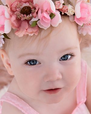 Adorable baby girl with blue eyes wearing a pink floral crown, looking at camera with a sweet expression.