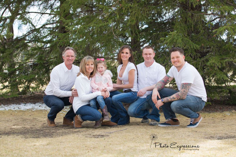 Casual family photo session with everyone sitting together on the ground near pine trees.
