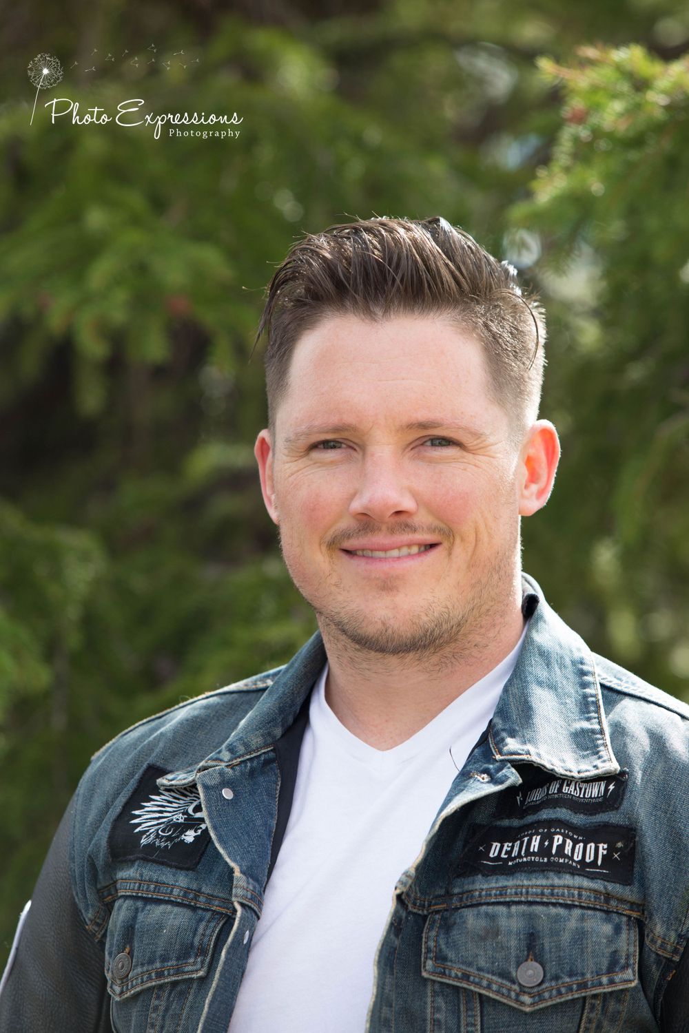 Portrait of person wearing white shirt and denim jacket against green foliage background.