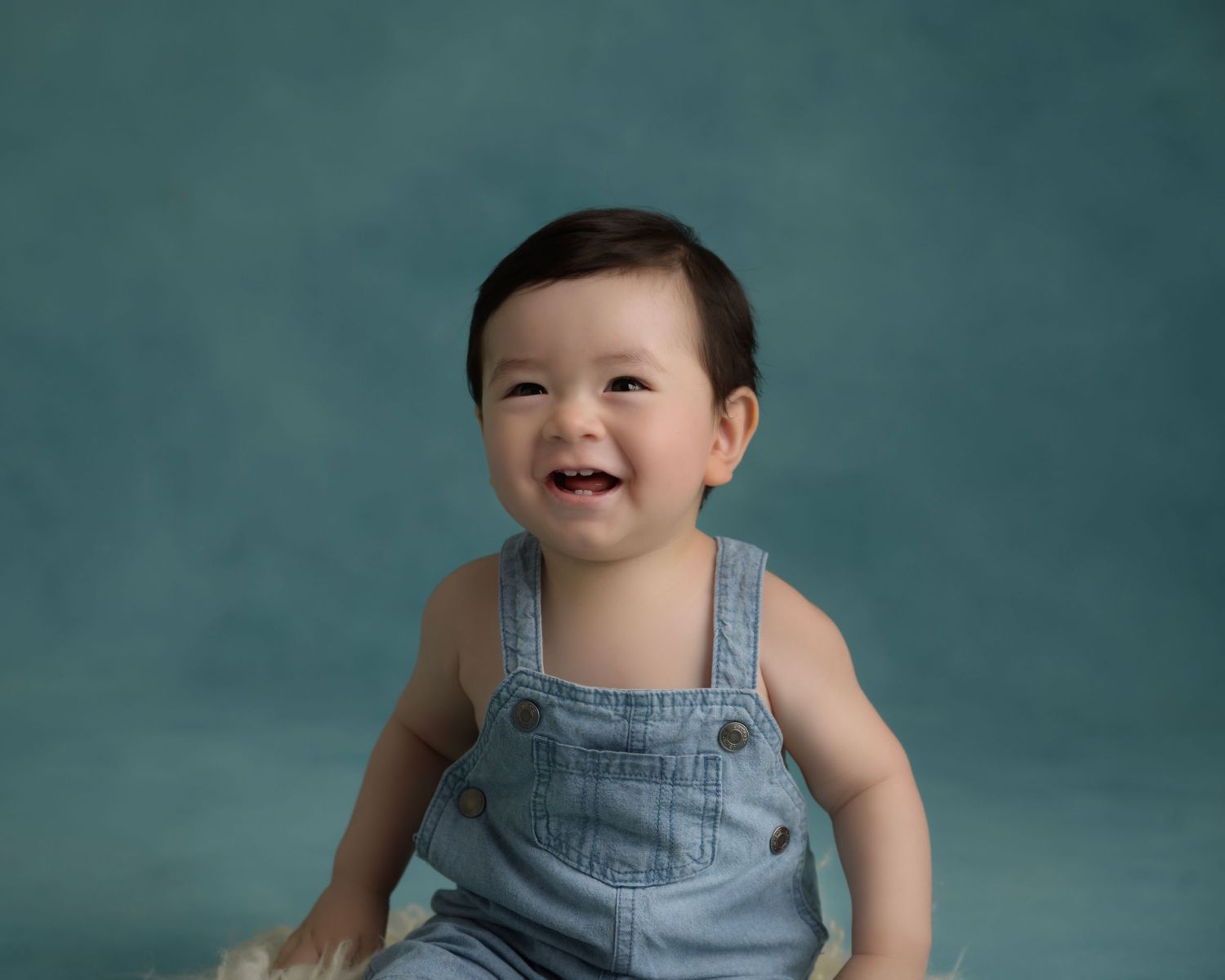 Laughing baby in blue denim overalls poses against teal studio background.