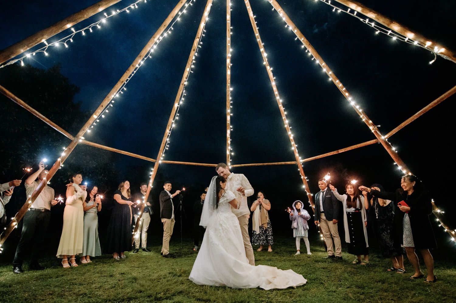 Wedding party gathers under illuminated tent poles at night during outdoor reception celebration.