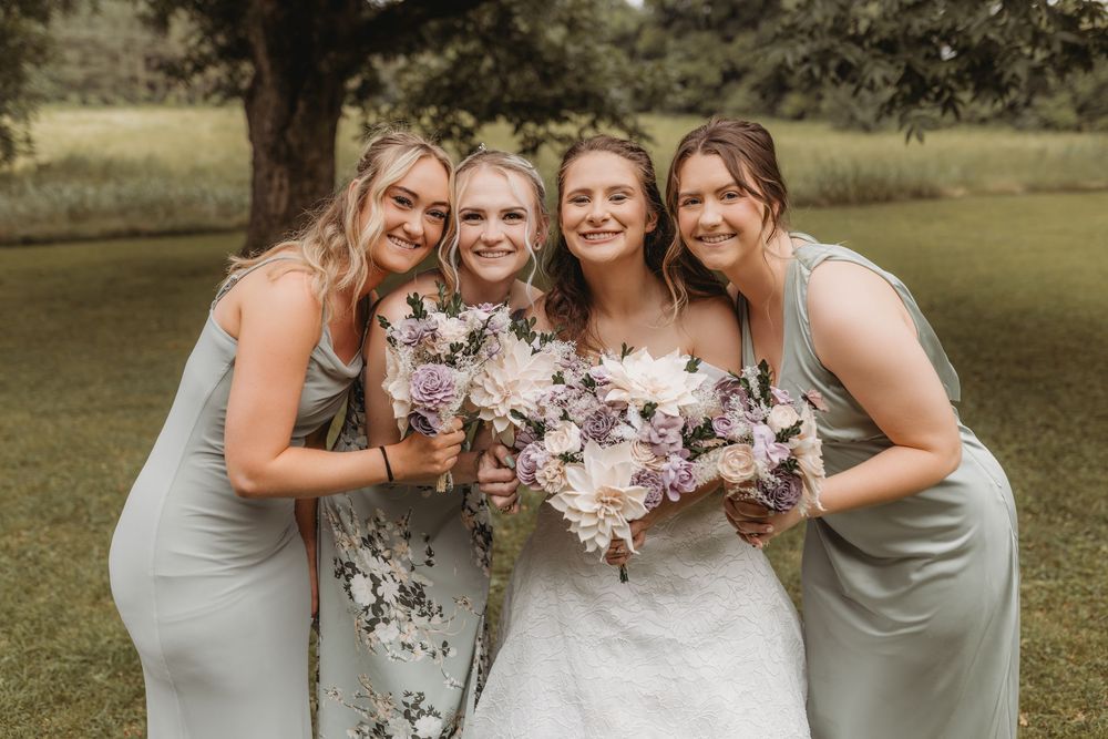 Bride with three bridesmaids in sage green dresses holding purple and white floral bouquets outdoors.